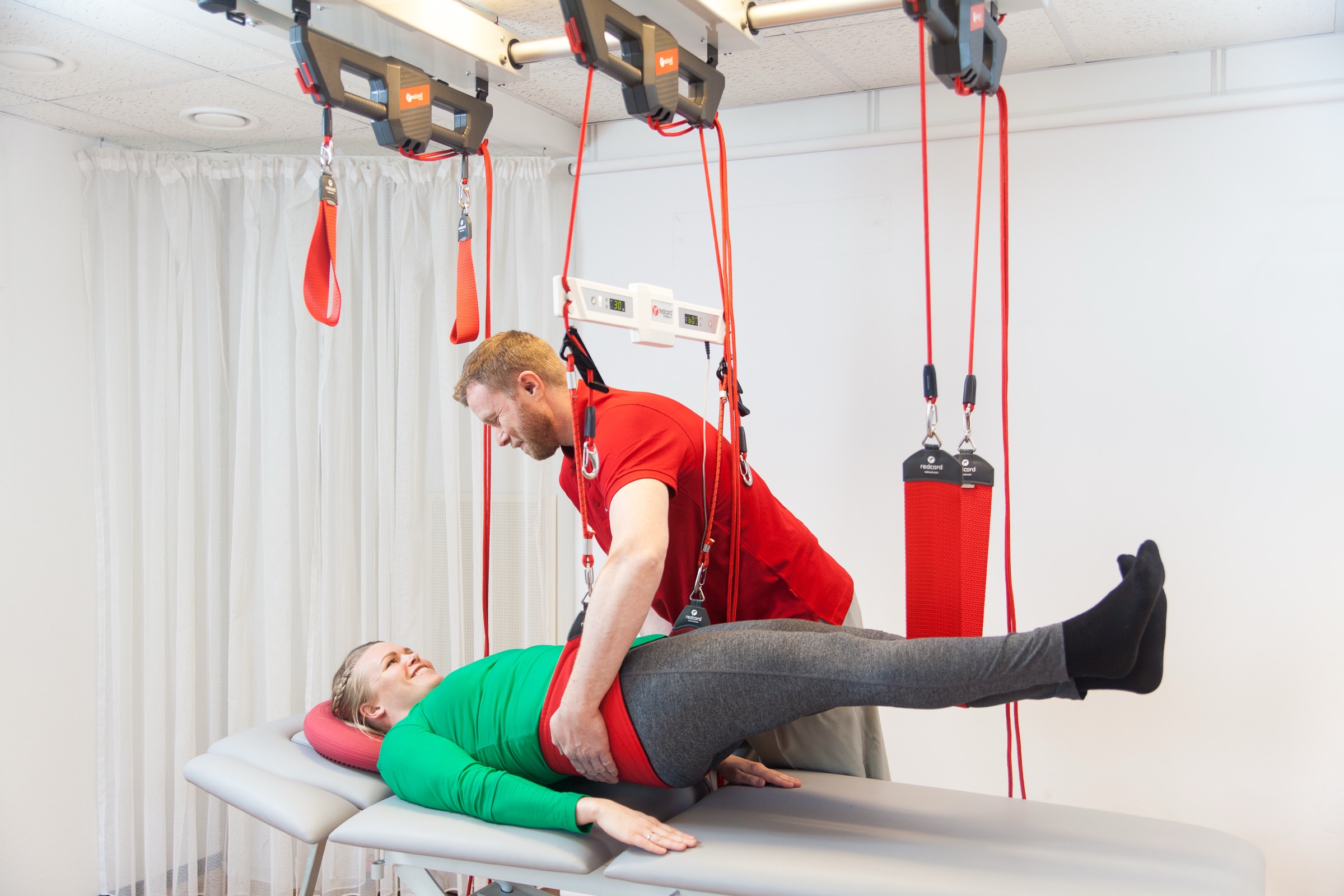 A practitioner supporting a patient through a supine leg exercise on a fully rigged Redcord Workstation